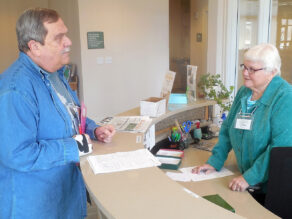 Sister Carol stands behind a reception desk and is talking with a reception volunteer.