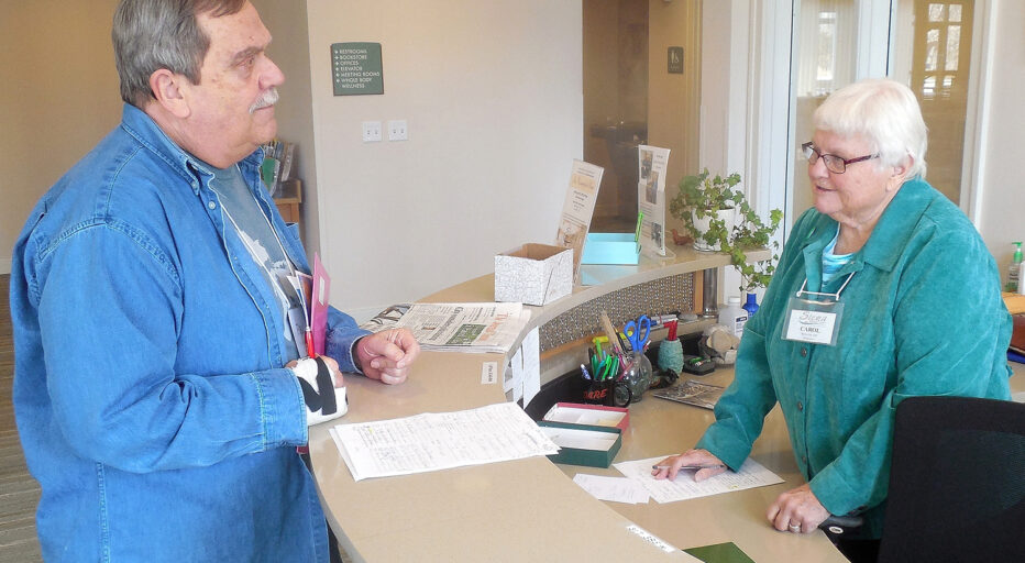 Sister Carol stands behind a reception desk and is talking with a reception volunteer.